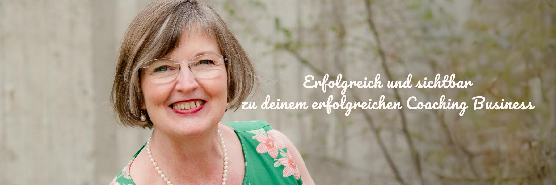 Erfolgreich sichtbar werden zum erfolgreichen Online Coach mit Elisabeth Engel spirituelle Businessmentorin. Portrait von Elisabeth Engel im grünen ärmellosen Kleid mit rosa Blumen vor grauer Wand mit grünen Sträuchern.